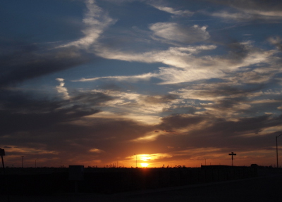 [The sun is just at the horizon and lights the clouds around it a bright yellow. Further away tp the sides the clouds are orange. The next level up in the sky the thick clouds are a medium grey while the swatches of clouds above that are very white against blue sky.]